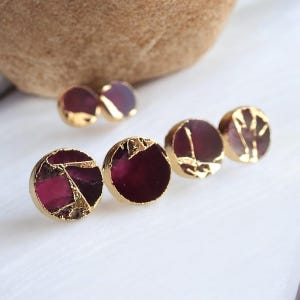 May include: Close-up of several pairs of gold-plated stud earrings. The earrings feature a round, dark red stone with gold accents. Some earrings have a cracked stone appearance, while others have gold leaf designs.
