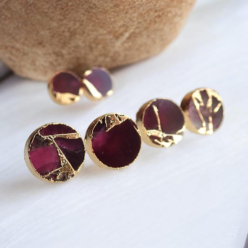 May include: Close-up of several pairs of gold-plated stud earrings. The earrings feature a round, dark red stone with gold accents. Some earrings have a cracked stone appearance, while others have gold leaf designs.