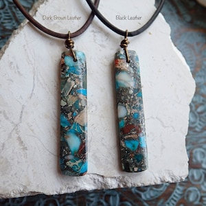 May include: Two necklaces with a blue and brown marbled stone pendant. The necklaces are on a white surface. The left necklace has a dark brown leather cord and the right necklace has a black leather cord. The text "Dark Brown Leather" is above the left necklace and the text "Black Leather" is above the right necklace.