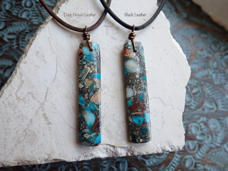 May include: Two necklaces with a blue and brown marbled stone pendant. The necklaces are on a white surface. The left necklace has a dark brown leather cord and the right necklace has a black leather cord. The text "Dark Brown Leather" is above the left necklace and the text "Black Leather" is above the right necklace.