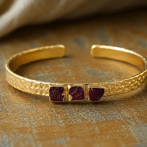 Pode incluir: Uma pulseira tipo bracelete dourada com textura de metal martelado e três pequenas pedras de corte bruto em vermelho escuro, fixadas horizontalmente no centro. A pulseira tem um design aberto para ajuste e um acabamento polido.