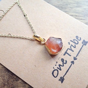 May include: A gold chain necklace with a raw carnelian stone pendant. The pendant is a natural, irregular shape and has a light orange color. The necklace is on a brown card with the text "One Tribe" written on it.