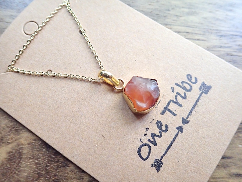 May include: A gold chain necklace with a raw carnelian stone pendant. The pendant is a natural, irregular shape and has a light orange color. The necklace is on a brown card with the text "One Tribe" written on it.