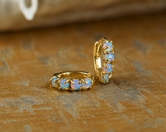 May include: A pair of gold hoop earrings featuring a row of small, round, iridescent opals set in prongs. One hoop is lying flat while the other stands upright, showcasing the reflective, milky white and blue tones of the gemstones against the polished gold metal finish.