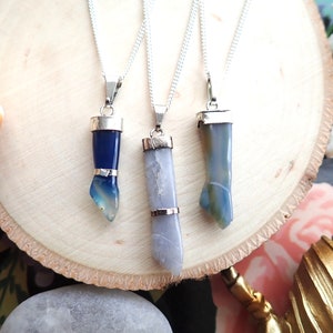 May include: Three silver necklaces with blue and grey agate stone pendants. The pendants are all different shapes and sizes. The necklaces are on a wooden surface.