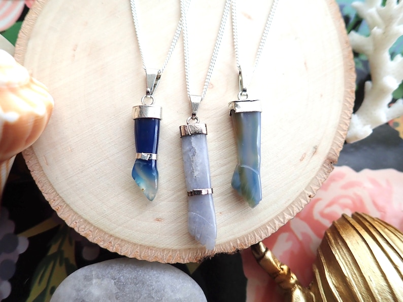 May include: Three silver necklaces with blue and grey agate stone pendants. The pendants are all different shapes and sizes. The necklaces are on a wooden surface.