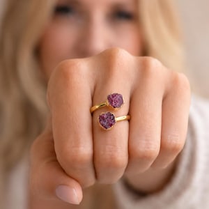 May include: A gold-toned bypass ring featuring two raw, deep magenta-colored stones. The stones have a textured, crystalline surface and are set at each end of the open band, which wraps slightly around the finger to display the two distinct gems.