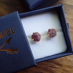 May include: A pair of raw, dusty rose-coloured gemstone stud earrings set in gold-toned prongs. The earrings are presented inside a dark navy blue cardboard jewellery box with the gold-embossed text "NE TRIBE" and a crossed arrow graphic on the lid.