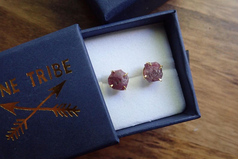 May include: A pair of raw, dusty rose-coloured gemstone stud earrings set in gold-toned prongs. The earrings are presented inside a dark navy blue cardboard jewellery box with the gold-embossed text "NE TRIBE" and a crossed arrow graphic on the lid.
