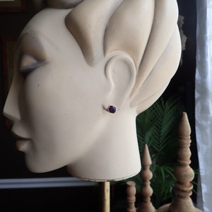 May include: A gold-colored stud earring with a small, dark purple gemstone is displayed on a mannequin head. The earring is set in a simple, classic design. The mannequin head is a light beige color.