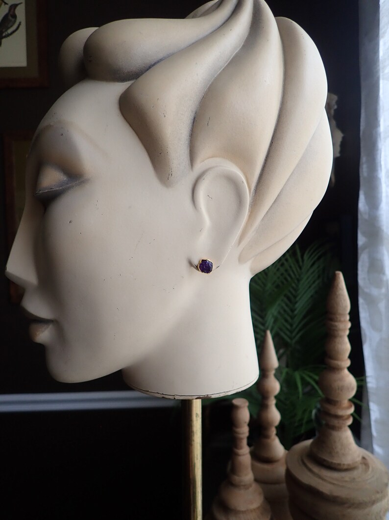 May include: A gold-colored stud earring with a small, dark purple gemstone is displayed on a mannequin head. The earring is set in a simple, classic design. The mannequin head is a light beige color.