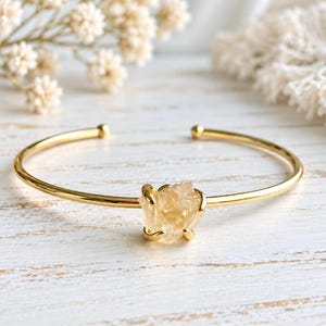 May include: A gold-toned open cuff bracelet featuring a single, raw, translucent yellow citrine crystal at the center. The crystal is held in place by four small gold-toned prongs, and the band has a smooth, polished finish with rounded ends.