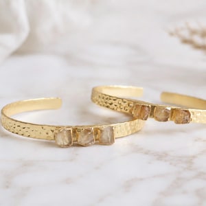 May include: Two gold-toned cuff bracelets featuring a hammered metal texture and three raw, translucent, light amber-colored stones set horizontally along the front of each open-ended band.