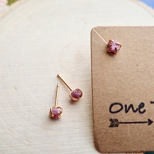 Puede incluir: Un par de pendientes de bot&oacute;n de oro con acentos de piedras preciosas crudas rosadas. Los pendientes se muestran sobre una superficie de madera con una tarjeta marr&oacute;n que dice "One Tribe".