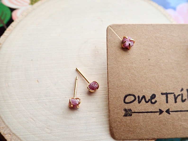 Puede incluir: Un par de pendientes de bot&oacute;n de oro con acentos de piedras preciosas crudas rosadas. Los pendientes se muestran sobre una superficie de madera con una tarjeta marr&oacute;n que dice "One Tribe".