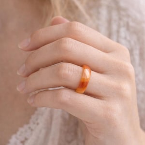 May include: A close-up of a ring made of orange and amber-colored material. The ring is on a finger, and the colors blend together in a marbled effect. The ring is a simple band, and the light reflects off the smooth surface.