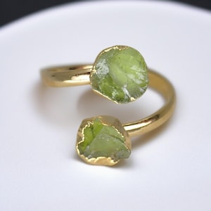 May include: A gold-colored ring featuring two raw, light green peridot gemstones. The peridot stones have a rough, natural texture and are set in gold-colored bezels. The ring's band curves around the finger, creating a unique, open design.