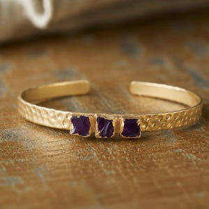 May include: A gold-toned metal cuff bracelet featuring a hammered texture along the band and three small, rough-cut, deep purple stones set horizontally in the center of the piece. The bracelet has an open back design for easy adjustment.