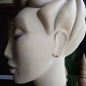 Puede incluir: Primer plano de una cabeza de maniqu&iacute; blanca con un peque&ntilde;o pendiente de piedra roja. El maniqu&iacute; tiene un peinado esculpido y est&aacute; mirando hacia la izquierda.