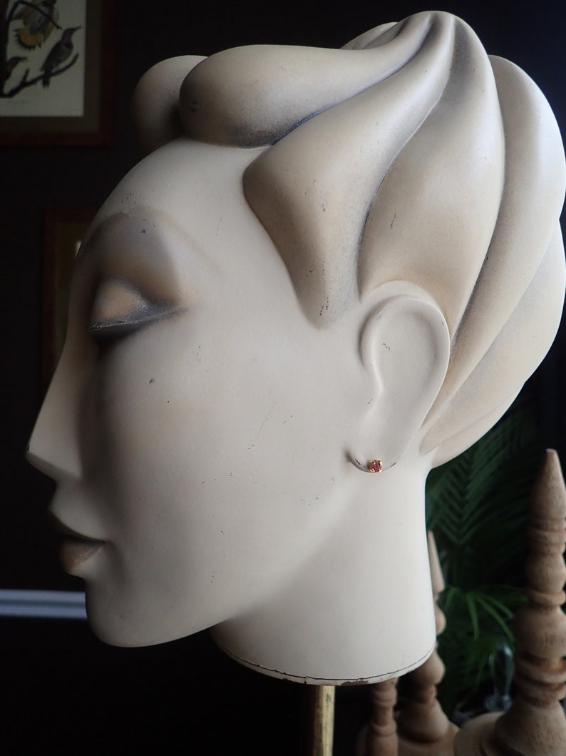 Puede incluir: Primer plano de una cabeza de maniqu&iacute; blanca con un peque&ntilde;o pendiente de piedra roja. El maniqu&iacute; tiene un peinado esculpido y est&aacute; mirando hacia la izquierda.