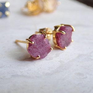 May include: A pair of stud earrings featuring raw, rough-cut pink gemstones set in gold-toned metal prongs. The stones have an irregular, textured surface and are secured by a four-prong setting. The earrings include a post back for attachment.