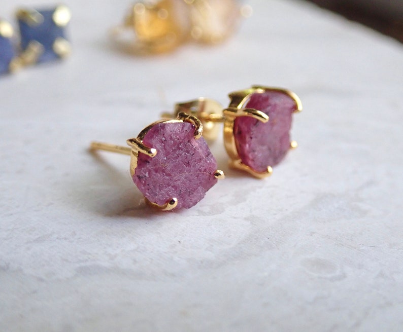 May include: A pair of stud earrings featuring raw, rough-cut pink gemstones set in gold-toned metal prongs. The stones have an irregular, textured surface and are secured by a four-prong setting. The earrings include a post back for attachment.