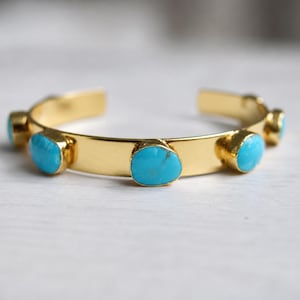 May include: A gold-colored cuff bracelet with six oval, robin's egg blue turquoise stones. The bracelet has a simple, open design. The stones are set in gold bezels. The bracelet is approximately 2.5 inches in diameter.