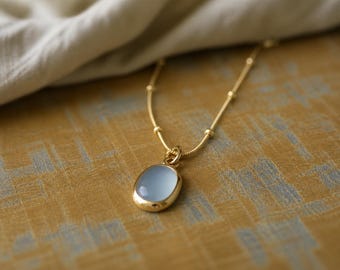May include: A gold-colored necklace with a light blue oval pendant. The pendant is framed in gold and hangs from a delicate chain. The background is a textured, golden-yellow surface.