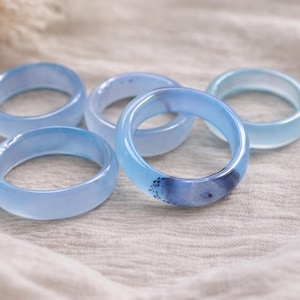 May include: Five light blue rings with varying shades of blue and a translucent appearance. The rings are made of a smooth material and are arranged on a neutral-colored fabric. The rings are a fashion accessory.