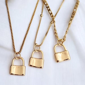 以下が含まれることがあります： 3つのゴールドカラーの南京錠ペンダントネックレス。各ネックレスは、繊細なチェーンに吊り下げられた小さな長方形の南京錠チャームが特徴です。チェーンには、ボックスチェーンやカーブチェーンなど、さまざまなスタイルがあります。背景は柔らかい白い生地です。