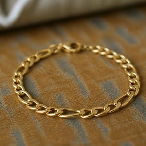 May include: A gold-colored Figaro chain bracelet. The bracelet features a pattern of three small circular links followed by one elongated oval link. The bracelet is displayed on a textured, neutral-toned surface.