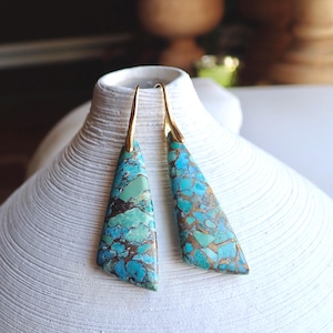 Puede incluir: Un par de pendientes colgantes de color turquesa y oro. Los pendientes presentan una piedra turquesa de forma triangular con motas doradas y un gancho dorado.