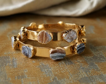 May include: Two gold-toned metal bangles featuring embedded, rough-cut, multi-coloured stone accents. The stones display layered patterns in shades of grey, brown, and white, set at intervals along the smooth, polished surface of each circular bracelet.