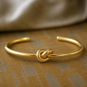 May include: A gold-colored cuff bracelet with a knot design in the center. The bracelet is made of a shiny, smooth metal and is displayed on a textured, golden-brown surface. The background features a soft, neutral-colored fabric.