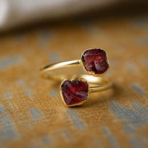 May include: A gold-colored ring with two raw, deep red gemstones. The gemstones are set in gold bezels, and the ring band curves around the finger. The ring is displayed on a textured, golden-brown surface.