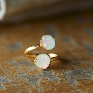 May include: A gold-toned bypass ring featuring two round, iridescent white stones with subtle flashes of color. The band is open at the front, allowing the two stones to sit opposite each other, creating a simple and clean design for the finger.