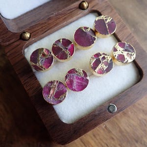 May include: Eight round stud earrings with a gold-coloured rim. The earrings feature a deep pink and gold-coloured stone. The earrings are displayed in a wooden box with a white interior.