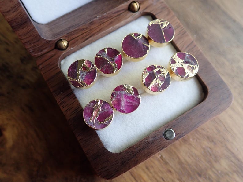 May include: Eight round stud earrings with a gold-coloured rim. The earrings feature a deep pink and gold-coloured stone. The earrings are displayed in a wooden box with a white interior.