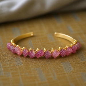 May include: A gold-toned cuff bracelet featuring a row of rough-cut, pink gemstones. The bracelet has a simple, open design with the gemstones secured by delicate gold wire. The background is a textured, golden-brown surface.