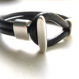 May include: Black leather bracelet with a silver-colored clasp. The bracelet features a loop design with a silver bar closure. The leather appears to be two strands. The bracelet is on a white surface.