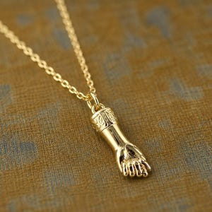 May include: A gold-colored pendant necklace featuring a detailed hand charm. The charm is suspended from a delicate chain. The hand pendant is intricately designed, showcasing the fingers and palm. The necklace is likely made of gold-toned metal.