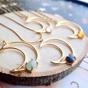 May include: Four gold crescent moon necklaces with different colored gemstones. The necklaces are displayed on a wooden surface. The gemstones are aquamarine, moonstone, sapphire, and citrine.