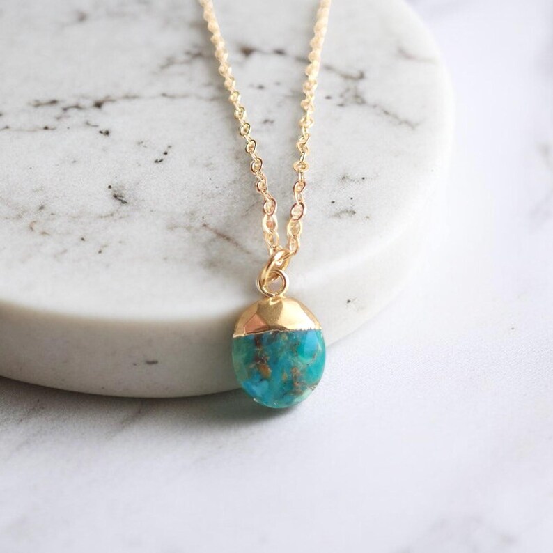 Peut inclure: Un collier &agrave; cha&icirc;ne dor&eacute;e avec un pendentif turquoise et or. Le pendentif est de forme ovale, avec un capuchon dor&eacute; et une pierre turquoise. Le collier est pr&eacute;sent&eacute; sur une surface en marbre blanc.