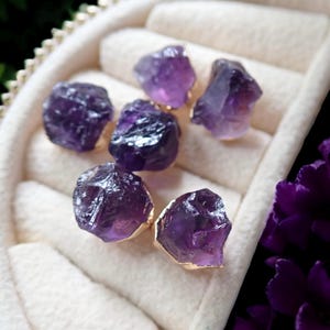 May include: Six raw amethyst gemstones with gold-colored edges are displayed in a white jewelry box. The gemstones are a deep purple color and have a rough, natural texture. The box has a soft, padded interior.