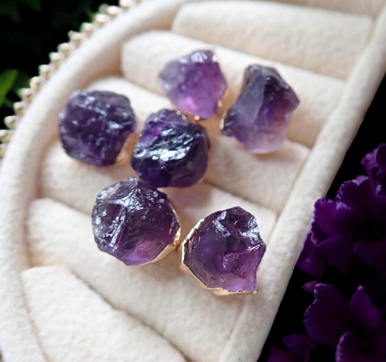 May include: Six raw amethyst gemstones with gold-colored edges are displayed in a white jewelry box. The gemstones are a deep purple color and have a rough, natural texture. The box has a soft, padded interior.