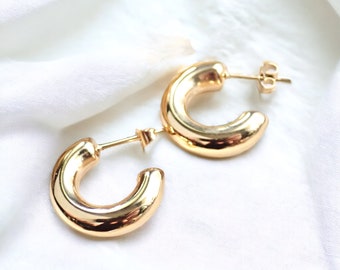 May include: A pair of gold hoop earrings with a thick, rounded design. The earrings are made of a shiny metal and have a simple, modern style.