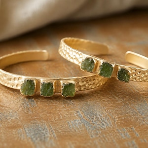 May include: Two gold-toned cuff bracelets featuring a hammered metal texture and three small, rough-cut green stones set in a row on the front of each band. The bracelets are open-ended, allowing for adjustment, and display a metallic finish.