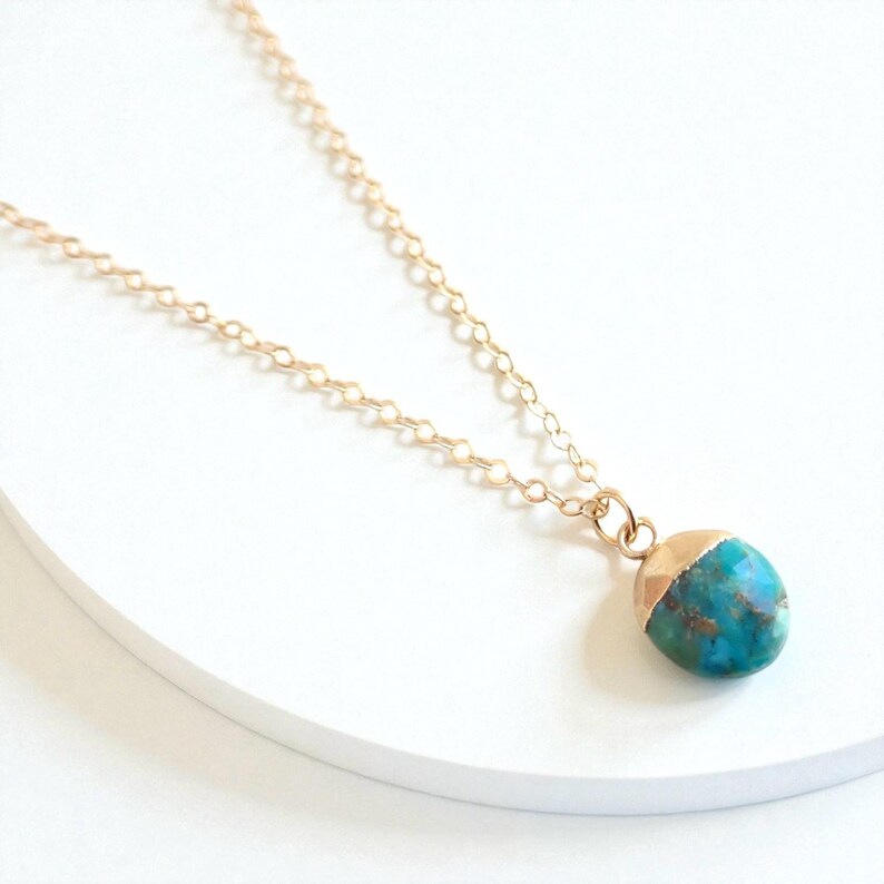 Peut inclure: Un collier &agrave; cha&icirc;ne dor&eacute;e avec un pendentif turquoise et or. Le pendentif est une pierre arrondie avec un capuchon dor&eacute;, pr&eacute;sentant un m&eacute;lange de teintes bleues et vertes. Le collier est pr&eacute;sent&eacute; sur une surface blanche.