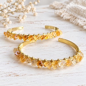 May include: Two gold-toned metal cuff bracelets featuring a row of raw, translucent yellow citrine crystal chips wired along the outer edge. Each bracelet has a circular band design with small metal beads separating the individual stones.
