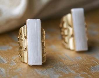 May include: Two gold rings, each featuring a rectangular white stone. The rings have a decorative cutout pattern. The rings are presented on a textured surface.